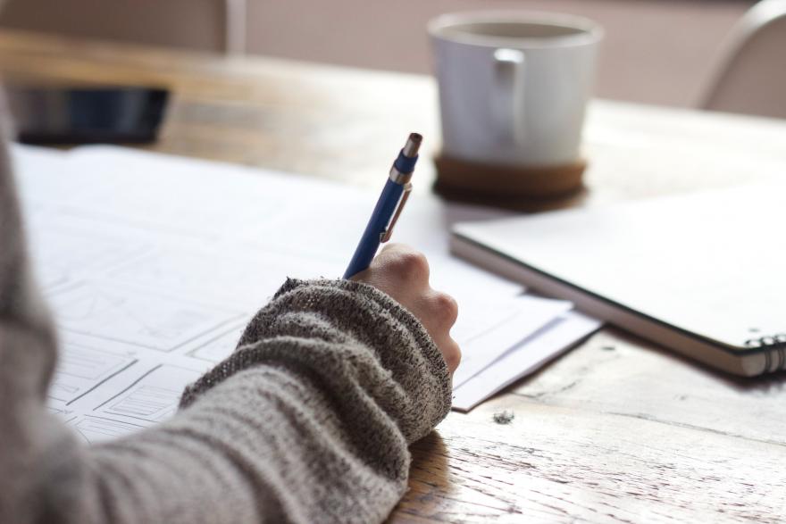 Mão segurando uma caneta sobre uma mesa com papeis, cadernos e xícara de café (foto: Green Chameleon/Unsplash)